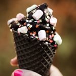 Trend cone of charcoal ice cream with Confetti and food powder, pink and blue fluffy sweet in women's arm. Blurred bokeh city background