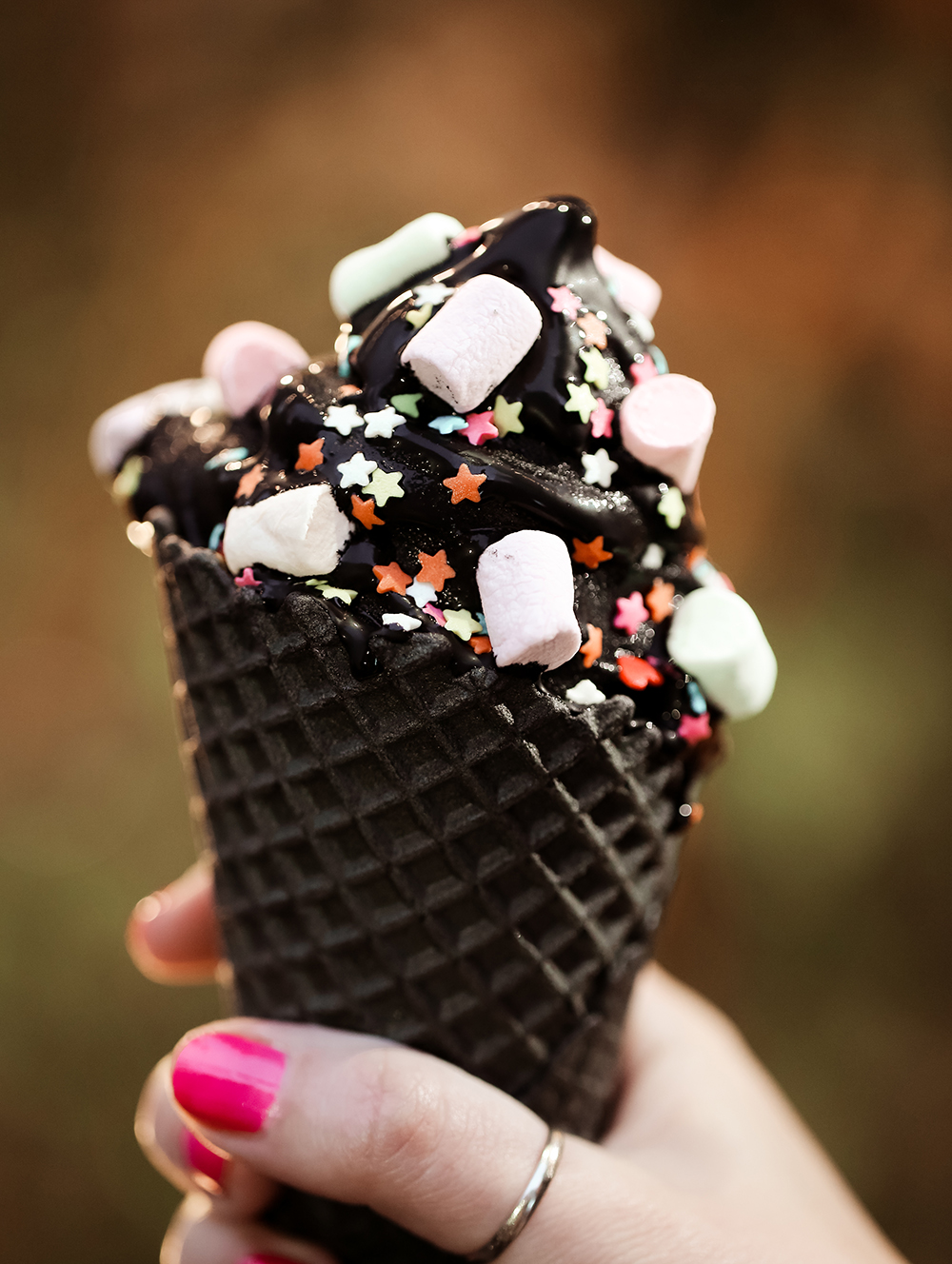 Trend cone of charcoal ice cream with Confetti and food powder, pink and blue fluffy sweet in women's arm. Blurred bokeh city background