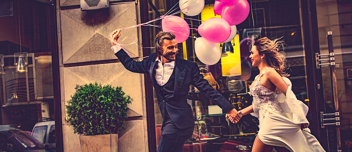 Lachendes Brautpaar mit Luftballons geht Hand in Hand die Straße entlang