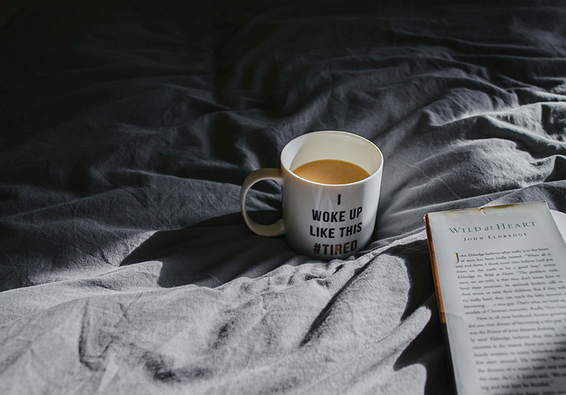 Kaffeetasse auf Bett grauem Bettlaken stehend mit einem Buch